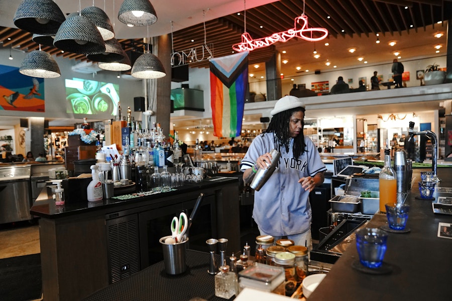 A bartender in a casual New York jersey mixes a cocktail behind a modern, vibrant bar decorated with a Pride flag.
