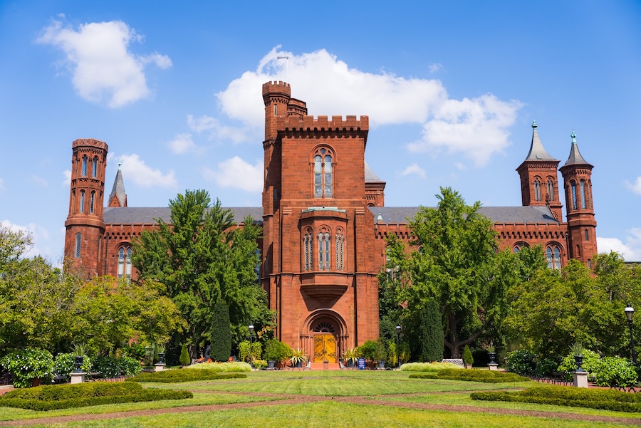 The Smithsonian Castle framed by greenery, seen from across the garden under a bright blue sky.