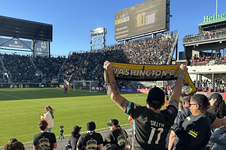 A Washington Spirit fan holds up a yellow scarf reading “Washington, DC” in a packed Audi Field stadium during a soccer match.