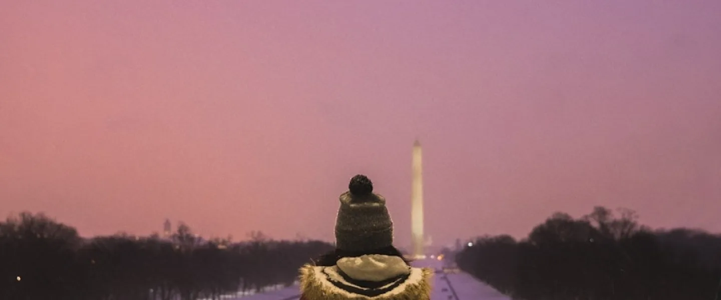 A person in a winter coat and hat stands at the Lincoln Memorial, looking out at the Washington Monument in the distance. The snowy landscape is illuminated by a soft, pinkish sky during twilight.