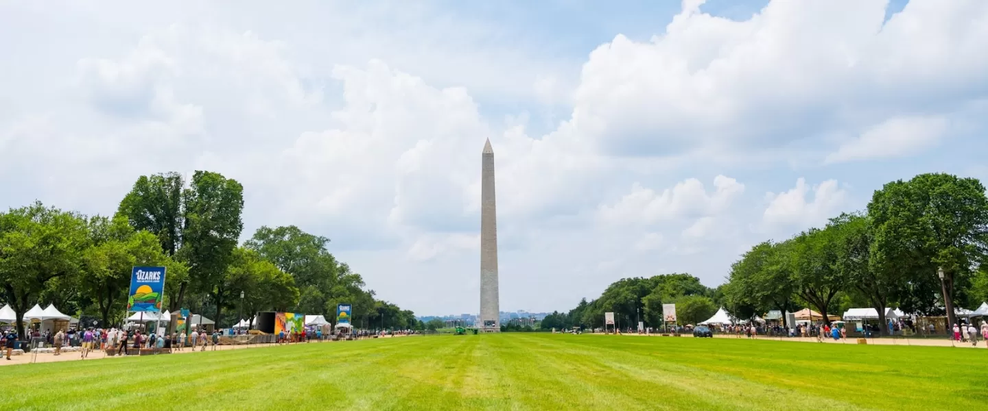 A view of the National Mall with the Washington Monument and a cloudy sky in the back. Festival-goers and tents are visible along the side of the green.