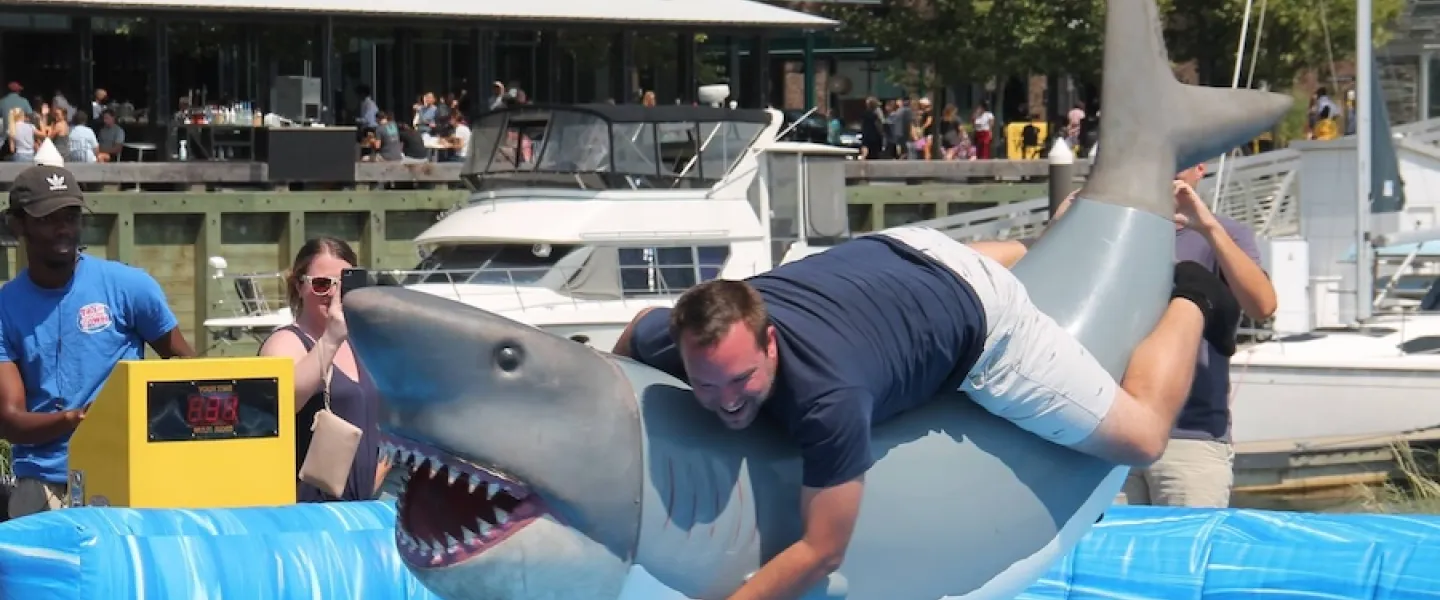 A man clings to a mechanical shark ride during an outdoor waterfront event.