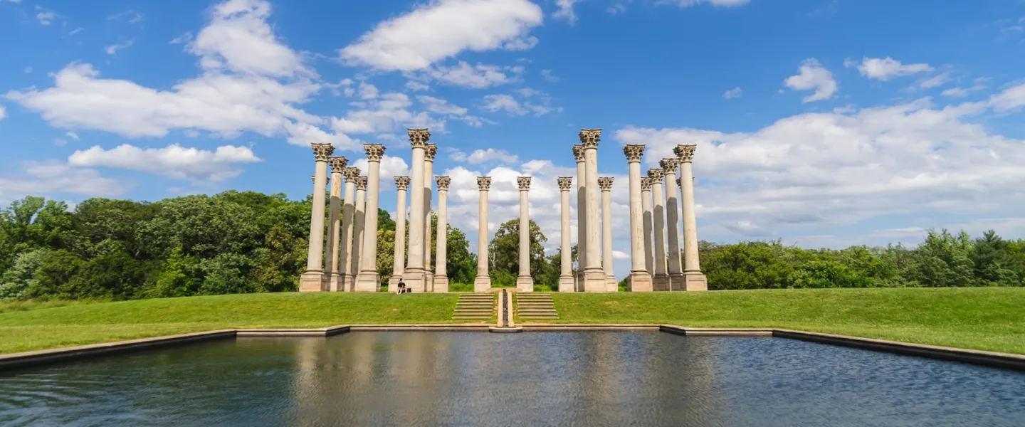 The National Capitol Columns stand tall on a grassy hill reflected in a pond at the U.S. National Arboretum under a bright blue sky.
