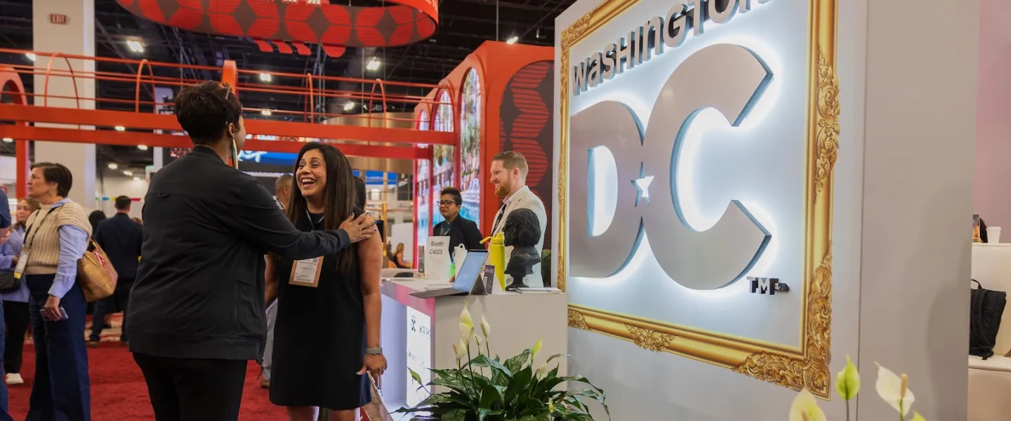 Attendees gather around a Washington, DC destination booth featuring illuminated signage and large digital displays at a travel trade show.