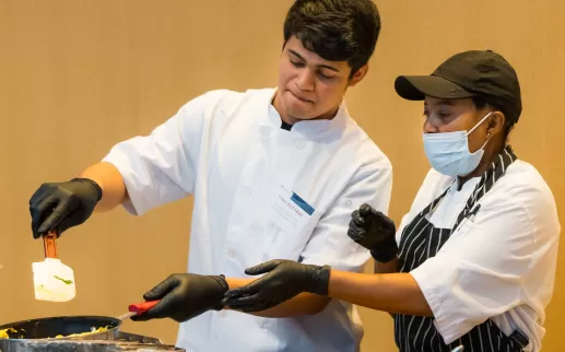 a young latino man in a chef's coat flips an omelet while an older black woman chef shows him the finer points of using his wrist to flip it 

