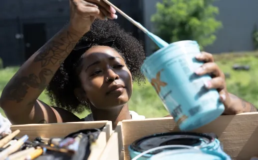 An artist dips a brush into a container of brightly colored paint.