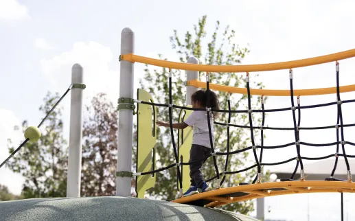 A young child climbs across a rope bridge on a colorful playground structure in Washington, DC.
