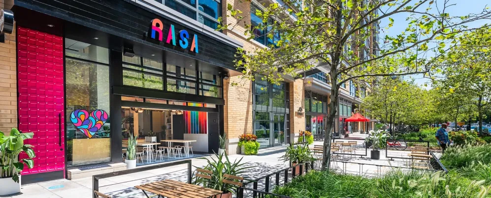 The exterior of Rasa restaurant in Mount Vernon Square features bright modern decor and a sunny outdoor patio with seating and greenery.